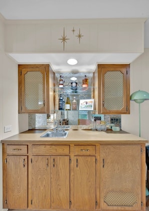 A kitchen with wooden cabinets, a glass display shelf, and a green floor lamp. - Retro Time Capsule · Mid-Century 3BR · Sleeps 9 (Shreveport)