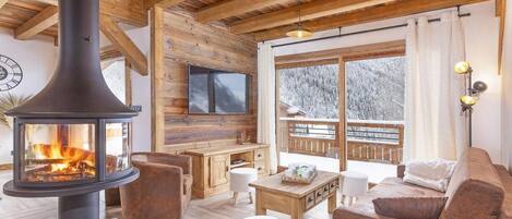 Een gezellige woonkamer met een houtkachel, comfortabele zitmogelijkheden, een televisie en een groot raam met uitzicht op een besneeerd landschap.