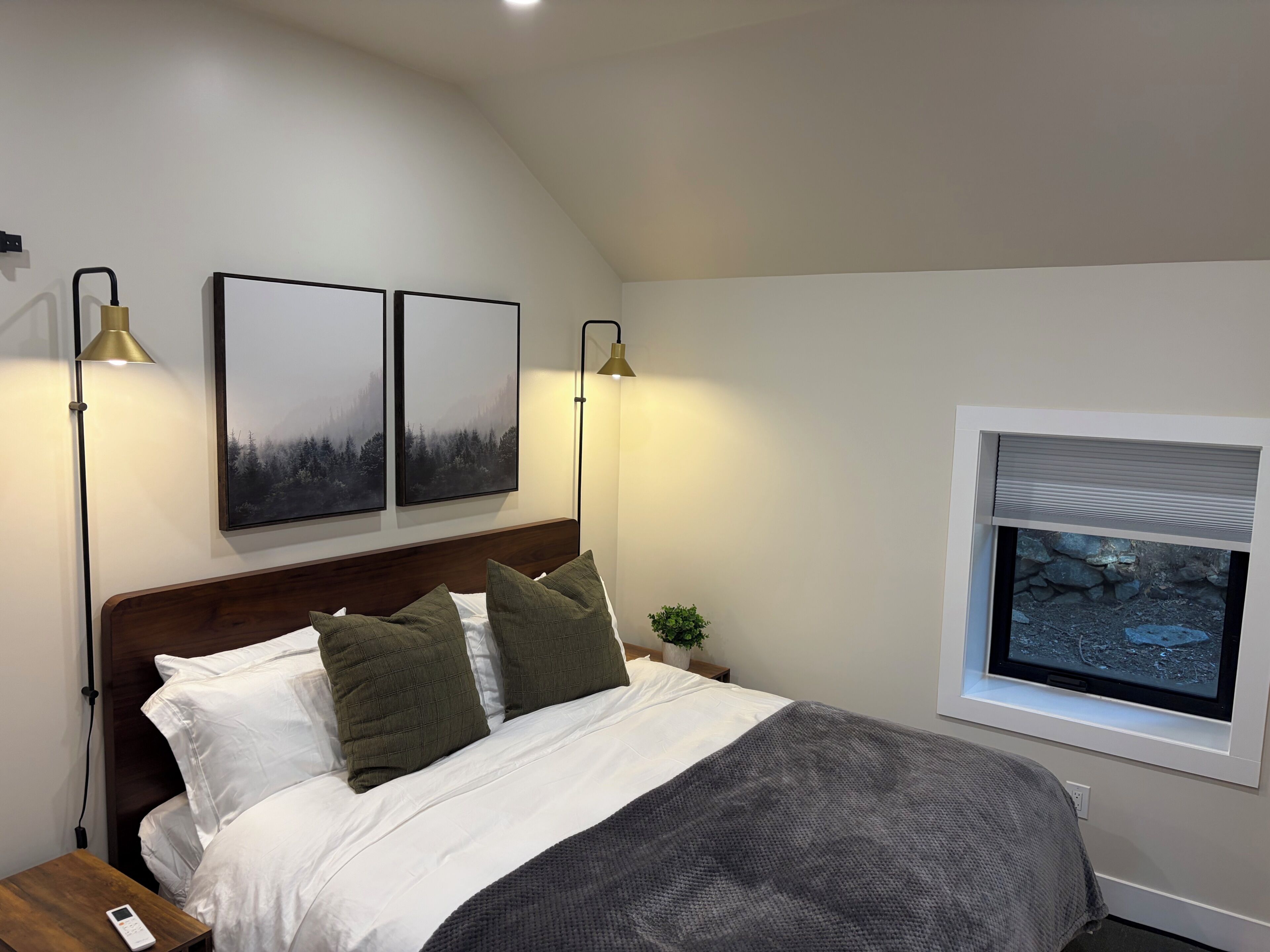 A bedroom with a bed, two wall-mounted lamps, a nightstand, a remote control, a window with a view, and two framed pictures.