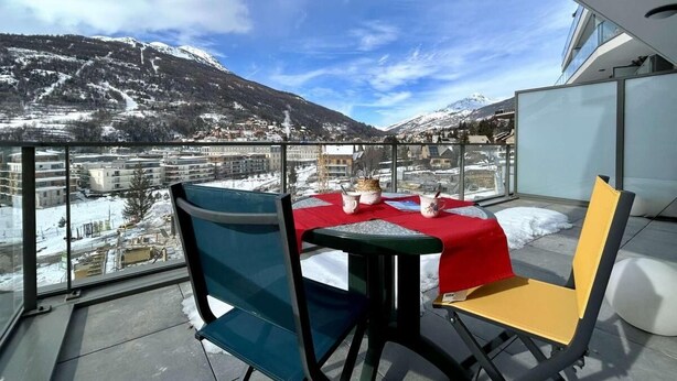 Un balcón con mesa y sillas, mantel rojo y vistas a una montaña nevada.