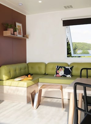 Ein modernes Wohnzimmer mit einem grünen Sofa, einem kleinen Tisch, einem Fenster mit Blick auf einen Wald und einem Regal mit Pflanzen.