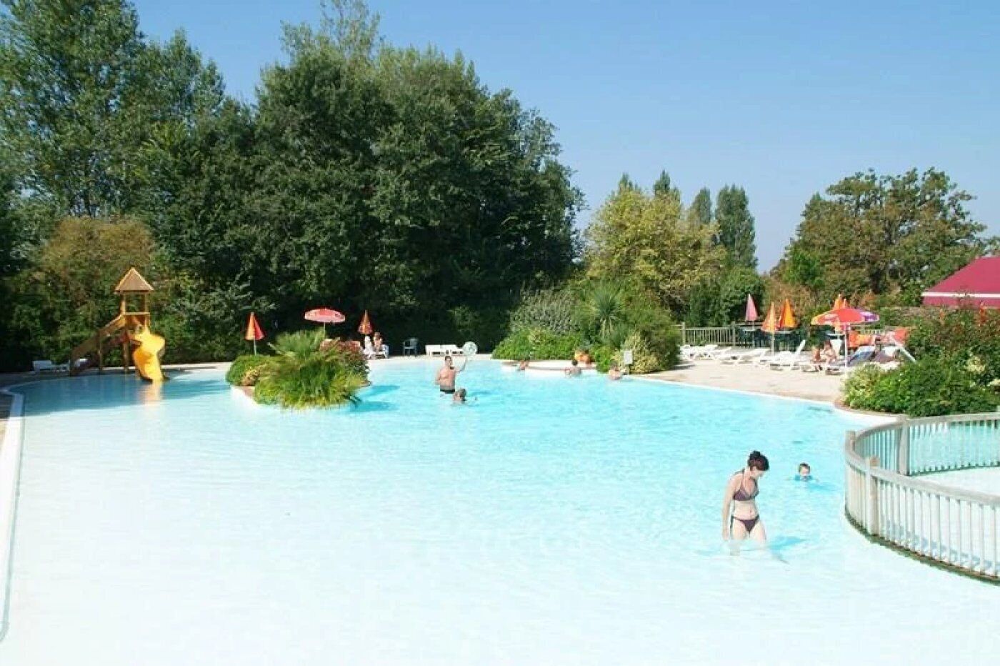 A swimming pool with a slide, surrounded by trees and umbrellas.