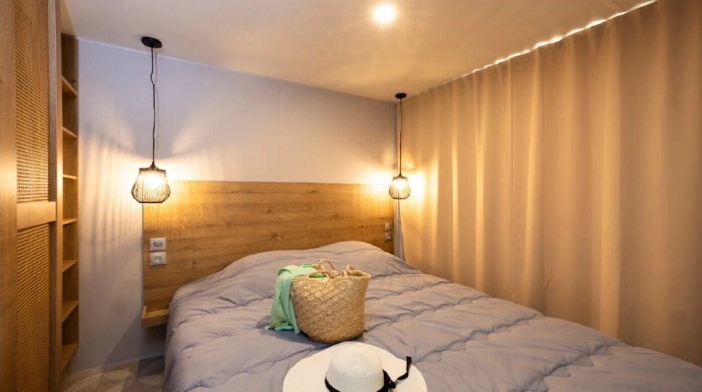 A bedroom with a wooden headboard, two wall-mounted lights, a bed with a grey quilt, a woven basket, and a white hat on the bed.