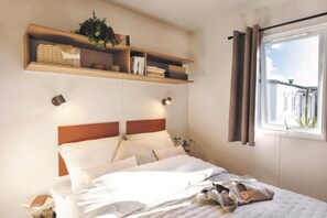 Ein Schlafzimmer mit Bett, Nachttischen, einem Fenster mit Vorhängen und einem Regal mit Büchern und einer Pflanze.