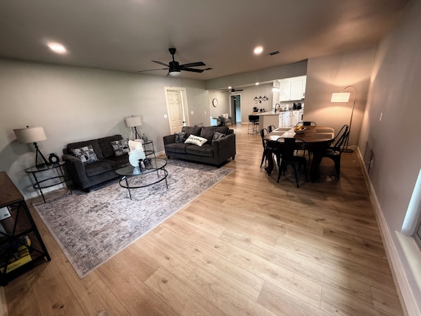 A spacious living room with a sofa, coffee table, and rug, leading to a kitchen and dining area with a table and chairs.