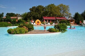 A swimming pool with a small island, lounge chairs, and a red-roofed building in the background. - Camping 4 étoiles - Parc aquatique - ccbfiad (La Romieu)