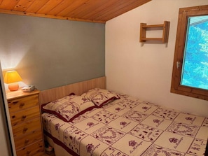 Ein Schlafzimmer mit einem hölzernen Kopfteil, einem Nachttisch mit Lampe, einer gesteppten Tagesdecke und einem Fenster mit Blick auf Bäume.