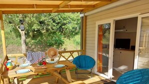 A wooden deck with a table set for two, a blue chair, and a covered grill area. - Camping 3 étoiles - Piscine  - ccbfiec (Les Moutiers-en-Retz)