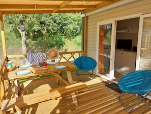 Una terraza de madera con una mesa preparada para dos, una silla azul y un área de parrilla cubierta.