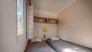 Ein Zimmer mit zwei Betten, einem fenster mit Vorhängen, einer Ablage mit einem gelben Gegenstand und einem Lichtschalter.