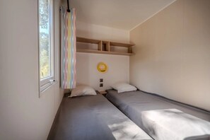 Ein Zimmer mit zwei Betten, einem fenster mit Vorhängen, einer Ablage mit einem gelben Gegenstand und einem Lichtschalter.