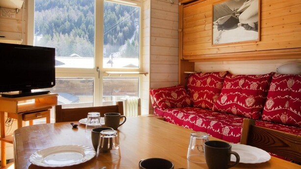Ein gemütliches Blockhäuschen-Interieur mit einem Holzespeisezimmer, einer Couch mit gemusterten Kissen, einem Fernseher und einem großen Fenster mit Blick auf einen Wald.