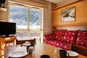 A cozy cabin interior with a wooden dining table, a sofa with patterned cushions, a television, and a large window with a view of a forest. - 250m from ski slopes, Ground floor, balcony, parking, tv, ski locker, 25m², La Norma (La Norma)