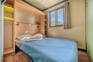Ein Einzelbett mit Kopfteil, Holzregale, ein Fenster mit Vorhängen und Blick ins Grüne.