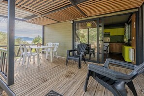 Eine überdachte Terrasse mit Tisch und Stühlen, eine Schiebetür zu einer Küche und Blick ins Grüne.