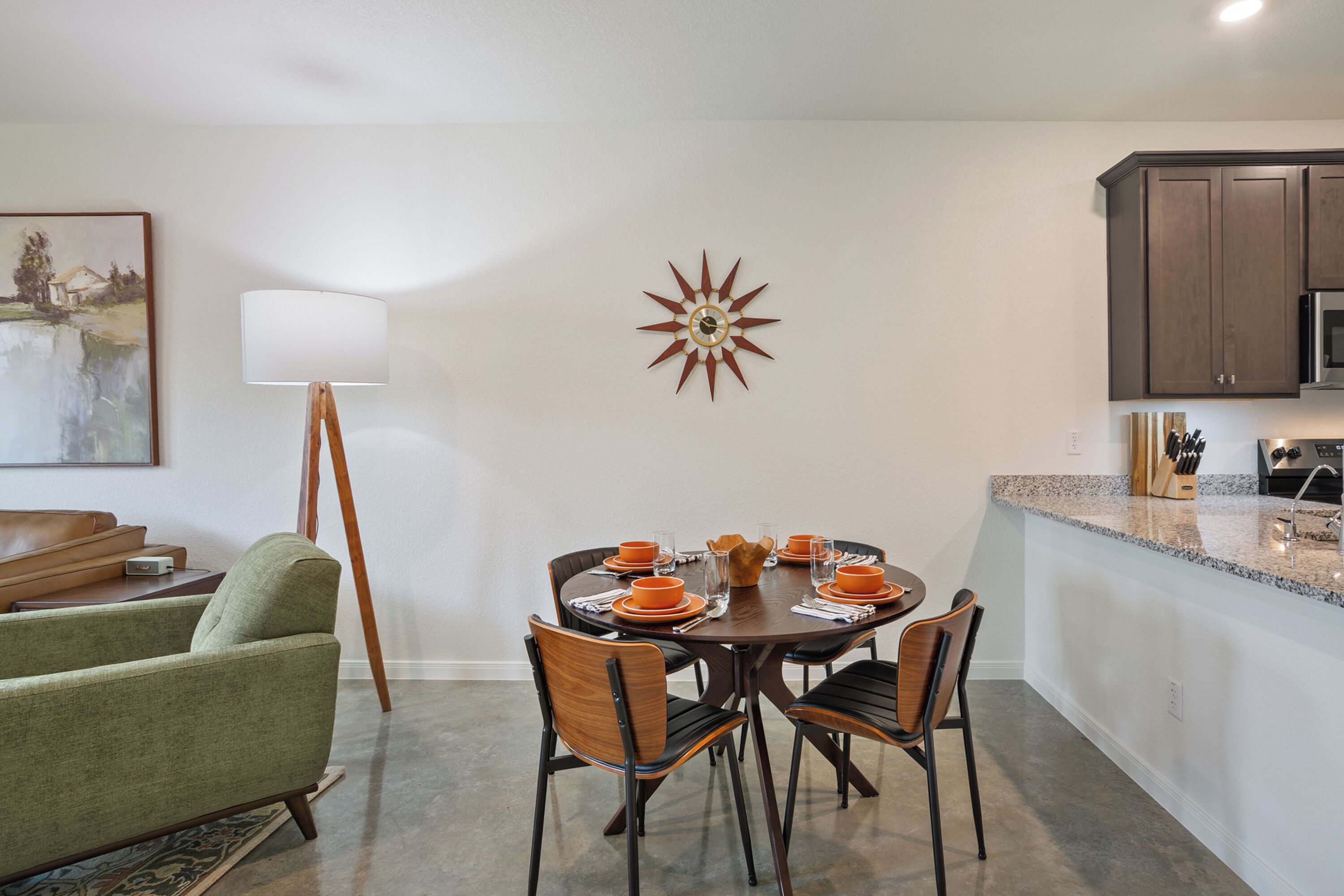 Un comedor con una mesa redonda dispuesta para cuatro personas, rodeada de sillas, una lámpara y un reloj en la pared.