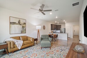 Ein Wohnzimmer mit Sofa, Sessel und einem gemusterten Teppich. Es gibt einen Deckenventilator, ein Gemälde und einen Essbereich mit Tisch und Stühlen.