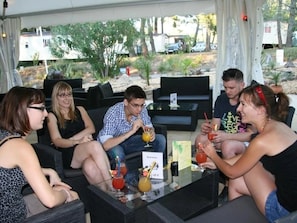 Um grupo de pessoas sentadas em uma mesa ao ar livre, com bebidas, cercado por outros assentos externos e vista para uma área residencial.