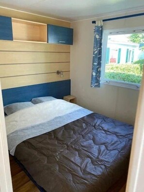 A small bedroom with a bed, a nightstand, a window with curtains, and a built-in shelf above the bed. - Camping 3 étoiles - Piscine  - ccbfhic (Rue)