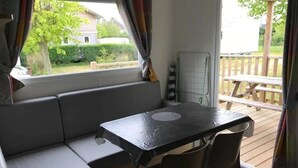 A compact living space with a sofa, a table, and chairs, a window with curtains, and a view of a deck outside. - Camping 3 étoiles - Piscine  - ccbfhid (Rue)
