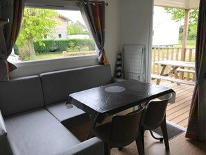 Um espaço de estar compacto com sofá, mesa, cadeiras, janela com cortinas e vista para um deck exterior.