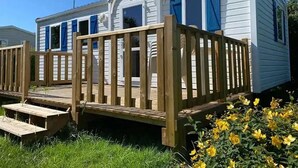 Une maison mobile dotée d’une terrasse en bois, d’un porche couvert et d’un petit jardin devant, agrémenté de fleurs jaunes.