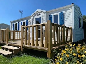A mobile home with a wooden deck, a covered porch, and a small front yard with yellow flowers. - Camping 3 étoiles - Piscine  - ccbfhie (Rue)