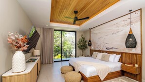 Une chambre moderne avec un grand lit, un ventilateur de plafond, une tête de lit en bois, une table de chevet, une commode, un vase avec des fleurs, une fenêtre avec des rideaux et une vue sur la verdure à l’extérieur.