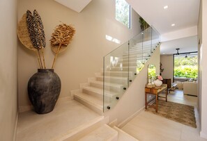 Eine moderne Treppe mit Glastrennwand, eine Vase mit dekorativen Blättern und Blick in einen Wohnbereich mit Sofa und Couchtisch.