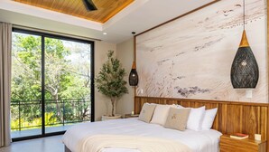 Une chambre avec un grand lit, une tête de lit en bois, une plante en pot, un luminaire suspendu et une vue sur la verdure par une porte coulissante.