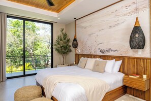 Ein Schlafzimmer mit einem großen Bett, einem hölzernen Kopfteil, einem Blumentopf, einer Pendelleuchte und Blick auf Grün durch eine Schiebetür.