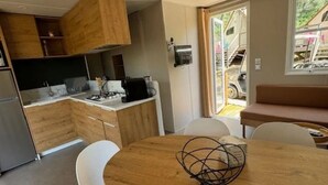 A modern kitchen with wooden cabinets, a dining table with white chairs, and a view of the outside through a glass door. - Camping 4 étoiles - Piscine  - ccbficf (Carqueiranne)