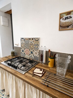 A kitchen with a gas stove, a patterned backsplash, a jar of spices, a yellow container with utensils, and a glass pitcher. - Fig Trees · Panoramic View and Pool (Saint Montan)