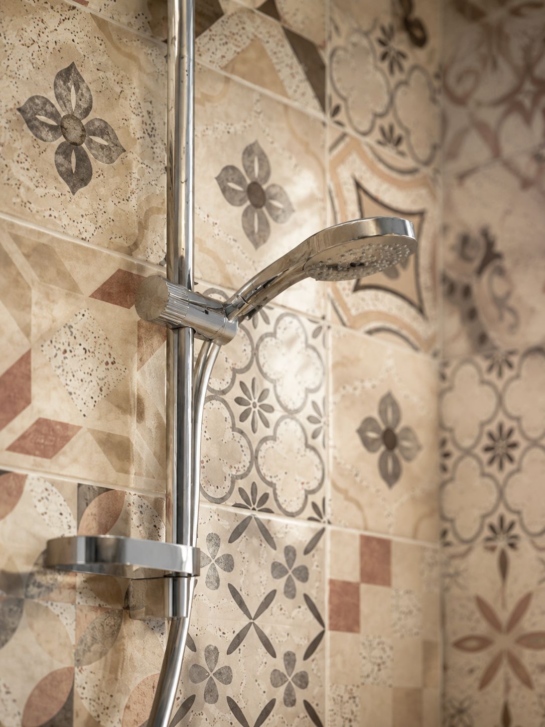 A shower with a chrome handle and a curved showerhead, set against a tiled wall with floral patterns.