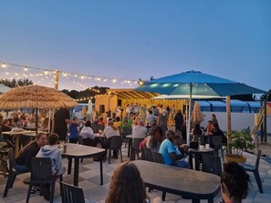 Ein Essbereich im Freien mit Tischen und Sonnenschirmen, umgeben von einem Gebäude mit einem Reetdach.