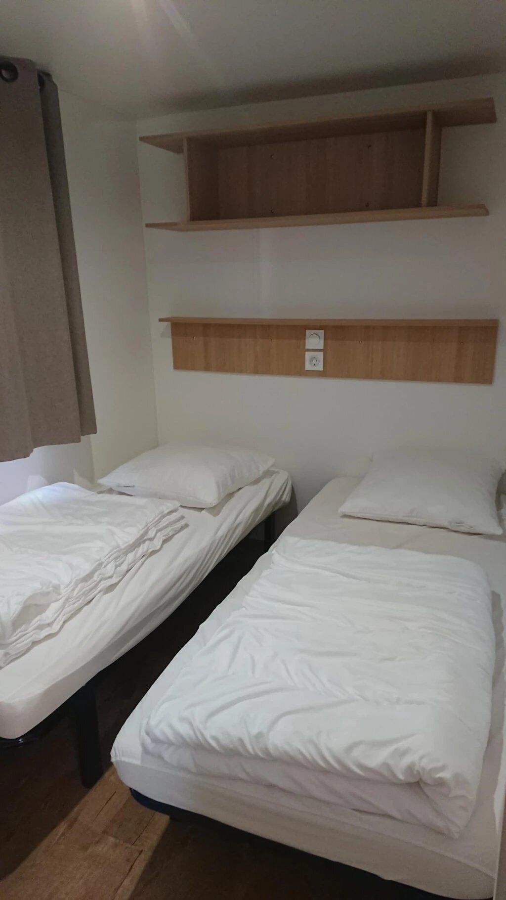 Une chambre avec deux lits simples, une étagère en bois au-dessus et une prise électrique sur le mur.