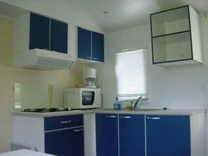 A kitchen with blue cabinets, a microwave, a stove, and a window with blinds. - Camping 3 étoiles - Piscine  - ccbgaae (Mens)