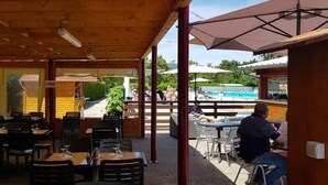 Et udendørs spiseområde med borde og stole, en pool i baggrunden og et overdækket siddeområde.