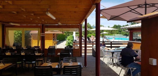 An outdoor dining area with tables and chairs, a pool in the background, and a covered seating area.