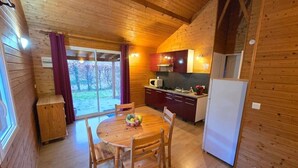 Interior de uma cabana de madeira com uma mesa de jantar, cadeiras, cozinha com armários e eletrodomésticos, e uma porta de vidro deslizante que dá para uma área externa.