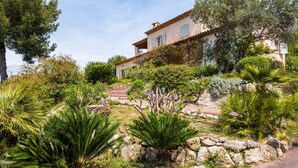Una villa de estilo mediterráneo con una escalera de piedra que lleva a la entrada, rodeada de abundante follaje y diversas plantas.