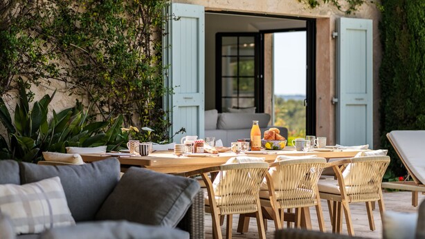 Un área de comedor al aire libre con sillas de mimbre, una mesa de madera y vista al interior a través de puertas abiertas.