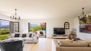 Amplia sala de estar con televisor grande, asientos cómodos y vistas a un jardín y montañas.
