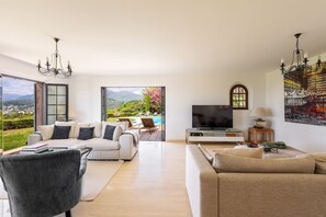 Ein geräumiges Wohnzimmer mit großem Fernseher, bequemen Sitzmöglichkeiten und Blick auf Garten und Berge.