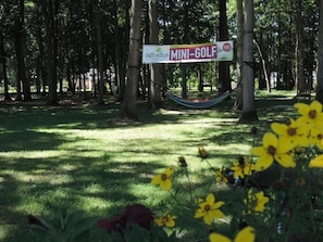 Schild für 'MINI-GOLF' in einem Park mit Bäumen und einer Hängematte.