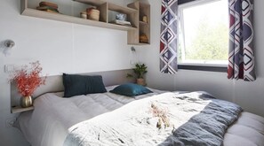 Ein Schlafzimmer mit Bett, einem Fenster mit gemusterten Vorhängen, einem Regal mit Gegenständen und einer Zimmerpflanze.