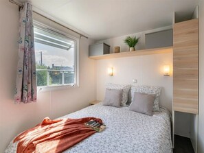 Ein Schlafzimmer mit Bett, einem Fenster mit Vorhängen, einem Regal mit Gegenständen und einer Wandleuchte.