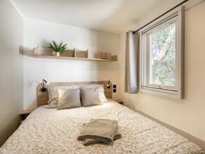 Ein Schlafzimmer mit Bett, Nachttisch, Fenster mit Vorhängen und einer Ablage mit Pflanze.