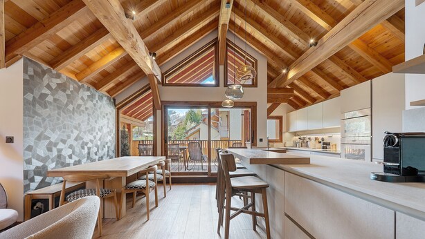 Una cocina moderna con vigas de madera, una zona de comedor con sillas y vistas a través de la puerta de cristal.