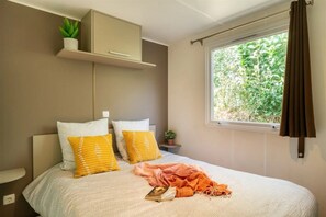 Ein Schlafzimmer mit einem Bett, Kissen, einem Fenster mit Vorhängen, einer Pflanze und einem Regal.